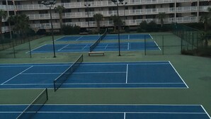 Canchas de tenis múltiples, rodeadas por una valla, y un edificio de varios pisos al fondo.