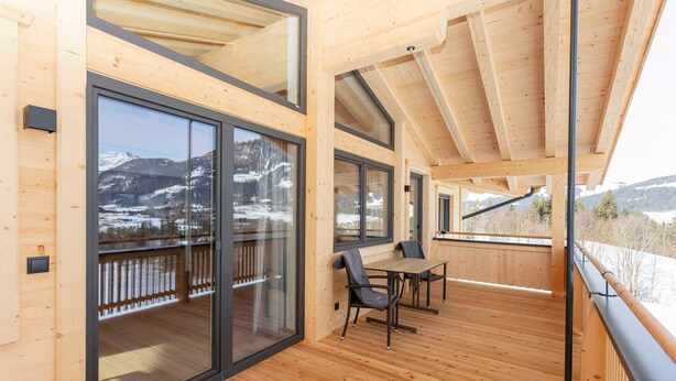 Eine Holzdachterrasse mit Tisch und Stühlen, großen Schiebeglastüren und Blick auf schneebedeckte Berge.