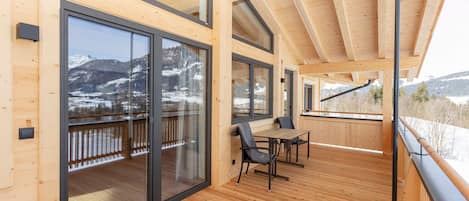 A wooden deck with a table and chairs, large sliding glass doors, and a view of snow-covered mountains.