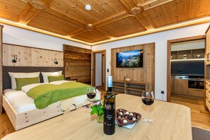 Techo de madera, cama con almohadas, televisor, mesa de comedor con vino y bombones, y una zona de cocina.