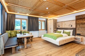 Un dormitorio con suelo de madera, una cama, un banco, una mesa y un ventanal con cortinas.