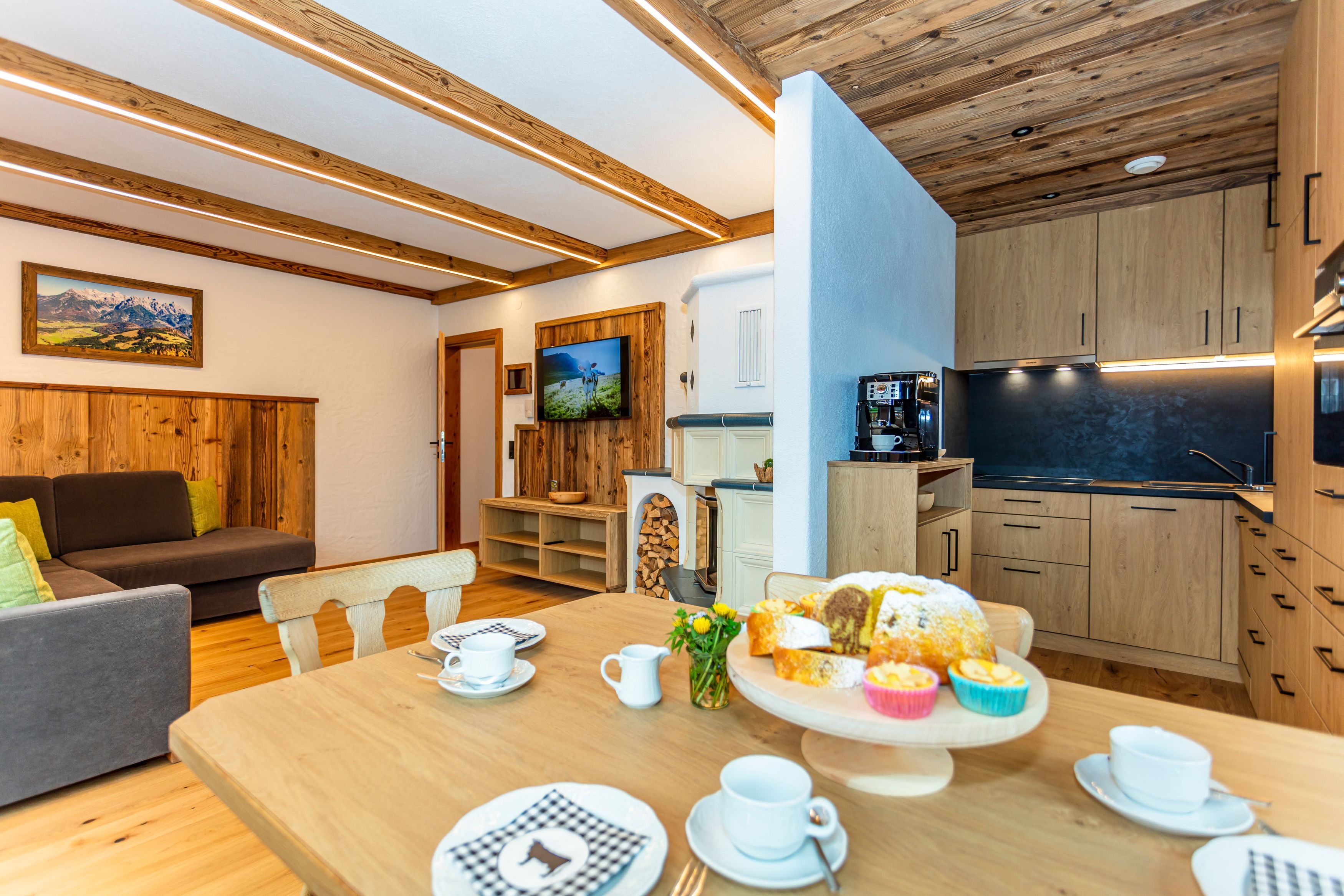 A modern kitchen with wooden cabinets, a dining area with a wooden table set for a meal, and a living room with a sofa and a TV.