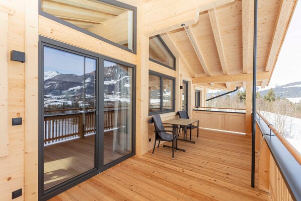 A wooden deck with a table and chairs, large sliding glass doors, and a view of snow-covered mountains.