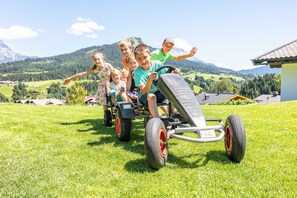 Un grupo de niños en un tractor de césped en un campo cubierto de hierba, con casas y montañas al fondo.