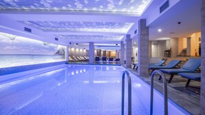 A modern indoor pool with a ceiling mural of the sea and clouds, lounge chairs, and a view of the ocean.