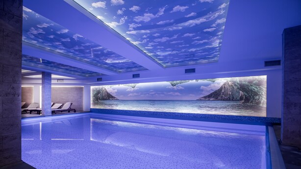 A modern indoor swimming pool with a ceiling mural of a sky and clouds, a large wall mural of a mountain and water, and lounge chairs along the poolside.