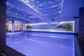 Ein moderner, überdachter Pool mit Deckenmalerei eines Himmels und Wolken, einer großen Wandmalerei von Bergen und Wasser sowie Liegestühlen am Pool.