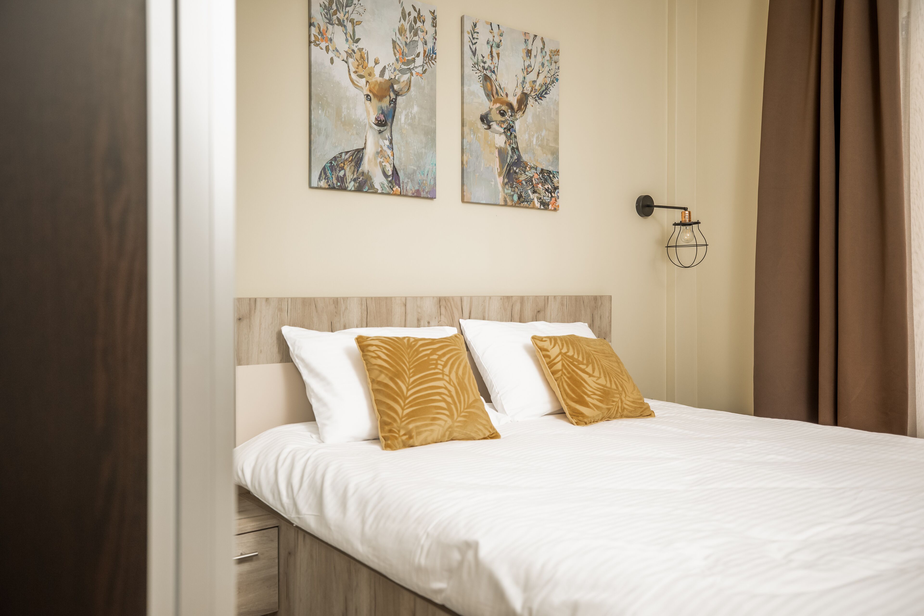 A neatly made bed with white and gold pillows, two wall-mounted paintings of deer, and a bedside lamp.