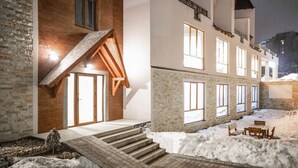 A building with a wooden staircase leading to a door, a snow-covered courtyard with a bench and table, and large windows on the upper floors.