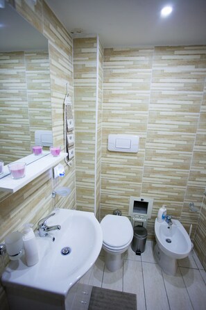 A modern bathroom with a large sink, toilet, bidet, and a mirror. There are toiletries on the sink and a trash bin.