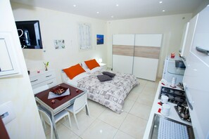 A compact hotel room with a bed, a desk, a chair, a kitchenette, and a wardrobe.