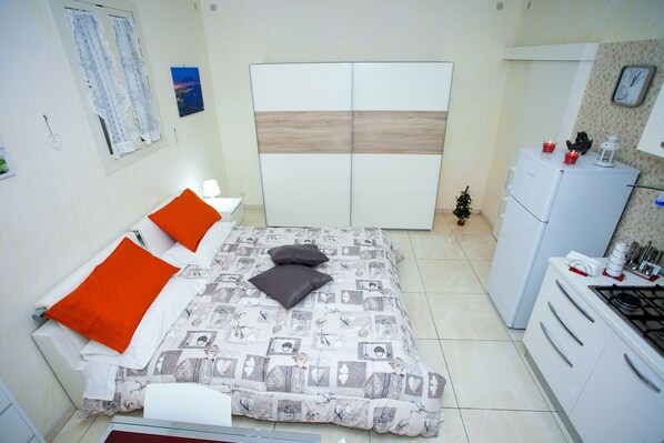 A bedroom with a bed, pillows, a nightstand, a wardrobe, a small Christmas tree, and a kitchenette.
