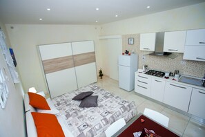 A bedroom with a bed, a small bedside table, and a nightstand. There is a kitchenette with a stove, sink, and cabinets.