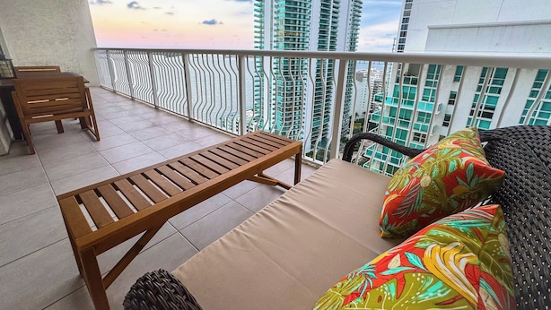 A balcony with a wooden table and a cushioned sofa with colorful pillows, overlooking tall buildings and a sunset. - Star Stay Miami / Orange@Brickell - King bed, Balcony & Free Parking (Miami)
