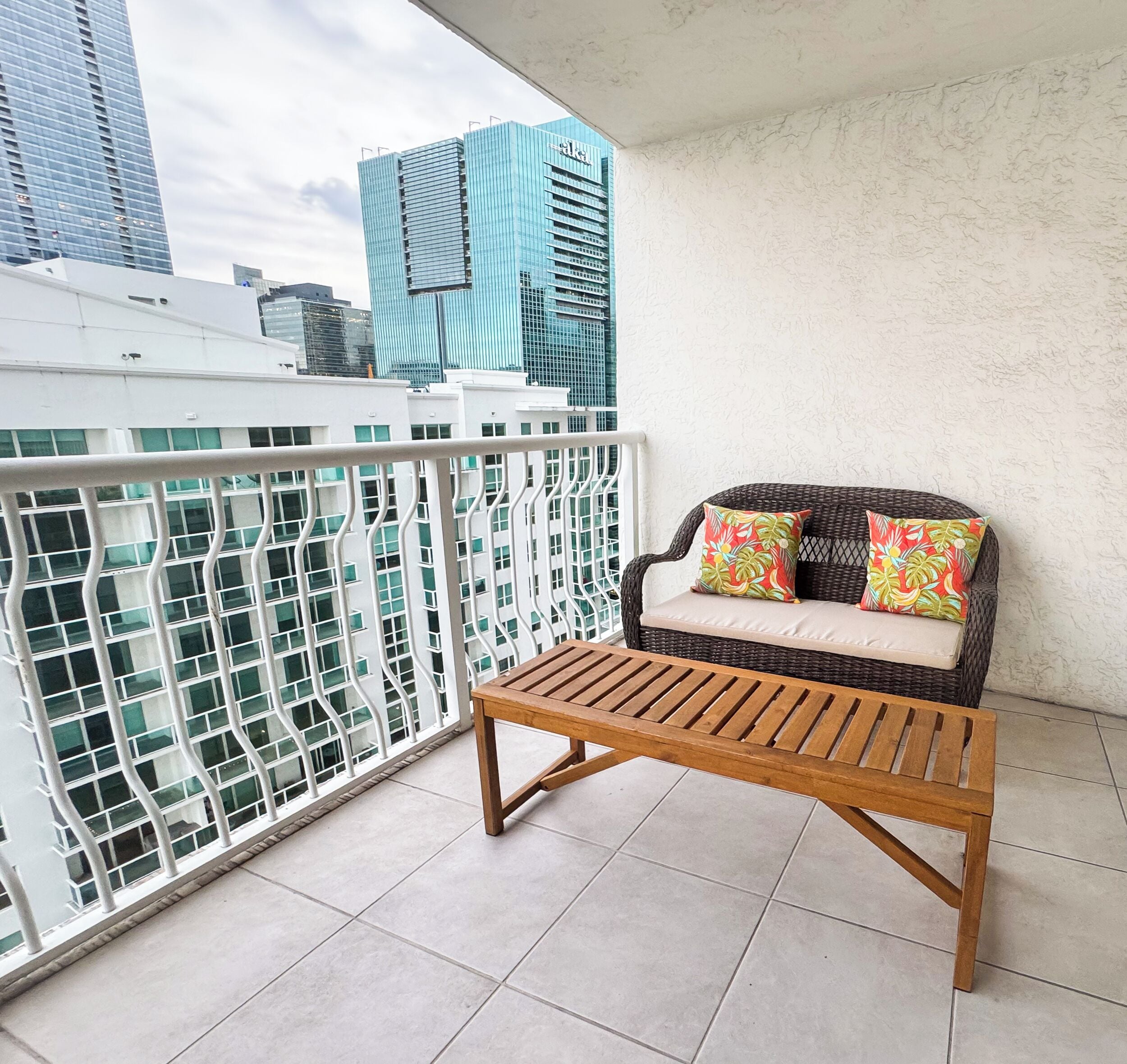 A balcony with a wicker sofa and a wooden table, a white railing, and a cityscape view.