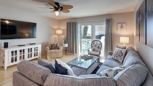 A living room with a large flat-screen TV, a ceiling fan, a white cabinet, a sofa, a coffee table, and a view of the outdoors through a sliding door. - Walton Dunes 7,  Beachfront, Private Beach.  2 Chair/1 Umbrella Set up Included. (Santa Rosa Beach)