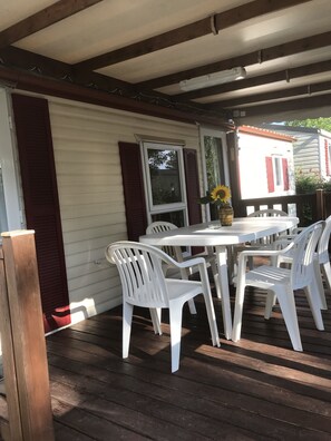 Un patio cubierto con una mesa y sillas blancas, una terraza de madera y una casa con contraventanas rojas.