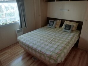 Une chambre à coucher avec un lit, une fenêtre avec des rideaux, un radiateur et une étagère fixée au mur.