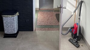 Un pasillo con una aspiradora, un cubo de basura y una estantería roja con varios objetos.