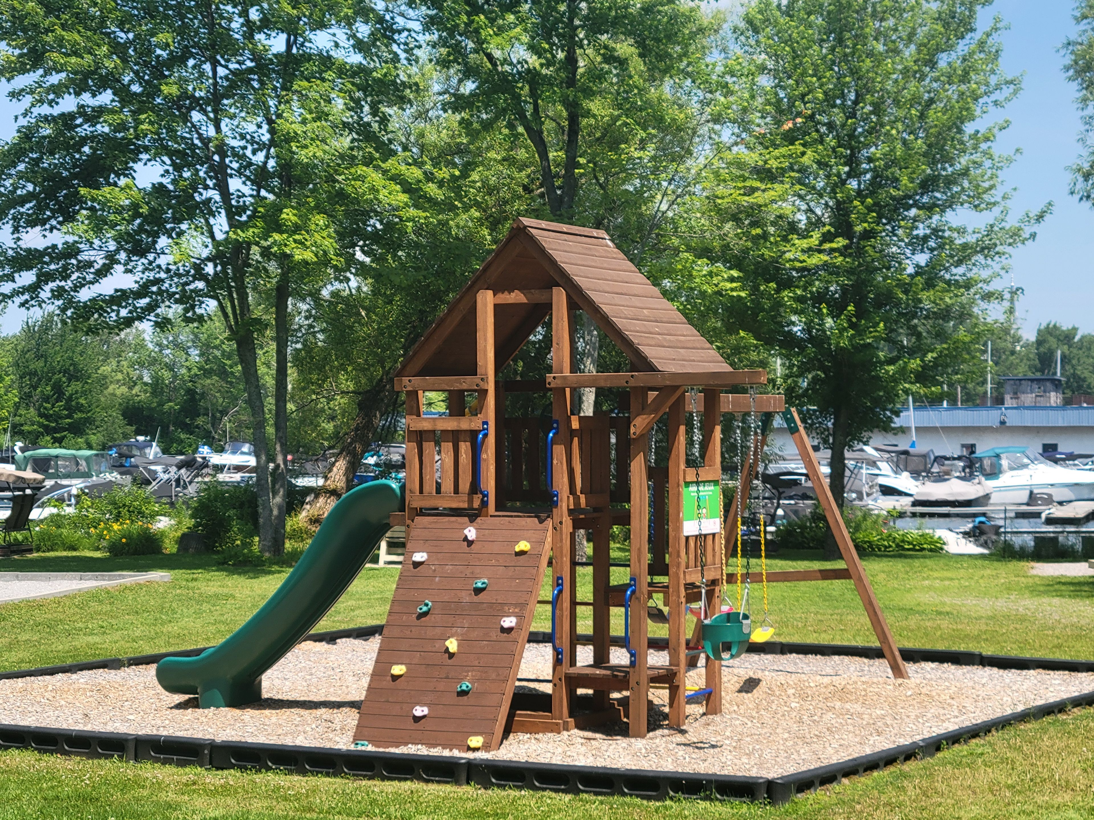 A wooden play structure with a slide and climbing wall, surrounded by a grassy area and trees.
