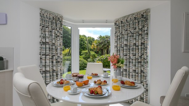 A breakfast table set with plates of food, cups, and a vase of flowers in a room with white walls and curtains. - Lamorna, Tregenna Estate, St Ives – Sleeps 4 – On-Site Parking for One Car (St Ives)