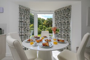A breakfast table set with plates of food, cups, and a vase of flowers in a room with white walls and curtains.