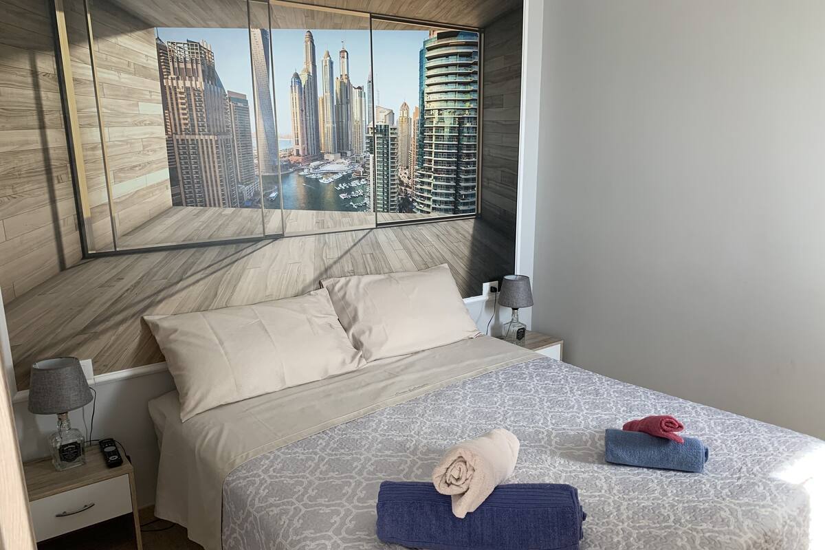 Een hotelkamer met een bed, twee nachtkastjes, een lamp en een stadsgezicht op de muur.