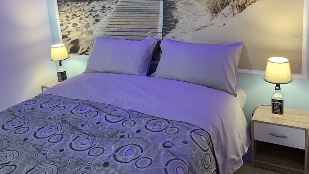 A bedroom with a bed, two bedside tables, a lamp, and a wall mural of a beach pathway. - The Lux (Adrano)