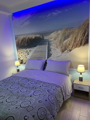 Ein Schlafzimmer mit einem Bett, zwei Nachttischen, einer Lampe und Wandgemälde eines Strandwegs.