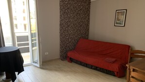 A room with a sliding glass door leading to a balcony, a red sofa, a wooden table, and a patterned curtain. - The Lux (Adrano)