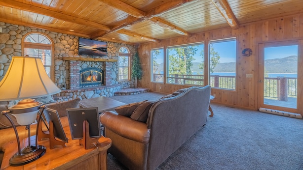 A cozy cabin interior with a stone fireplace, wooden ceiling beams, and large windows offering a view of the outdoors. - Valley View Cabin,Lake view, 6 bedrooms, Hot tub, walk distance to village (Big Bear Lake)