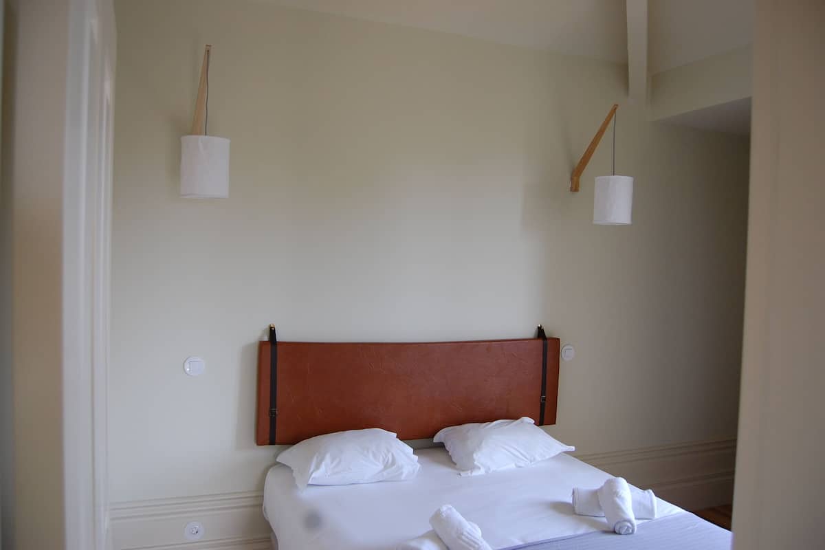 A neatly made bed with white linens and pillows, a wooden headboard, and two wall-mounted lamps.