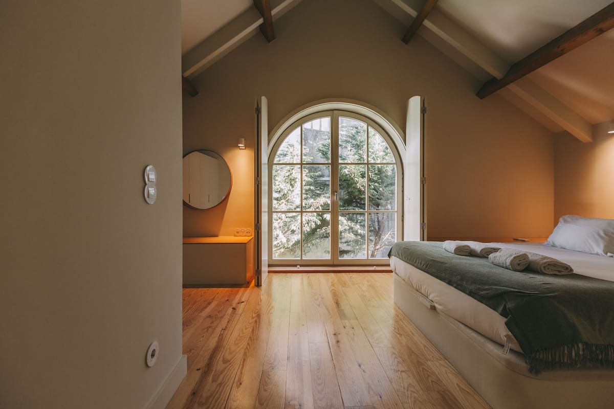 A bedroom with a large arched window, wooden flooring, a bed with a green blanket, and a round mirror on the wall.