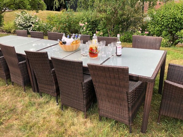 Un espacio de comedor al aire libre con sillas de mimbre y una mesa de vidrio decorada con botellas y un cuenco de fruta.