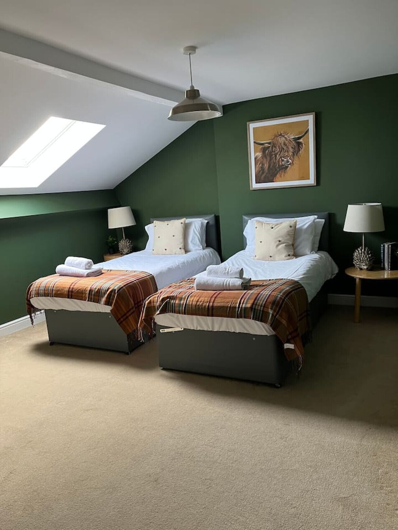 A room with two beds, a skylight, a painting of a yak, and a round table with a lamp.