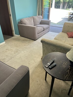 Un salón con un sofá color beige, una mesa redonda con dos mandos a distancia, y vistas a un patio exterior.