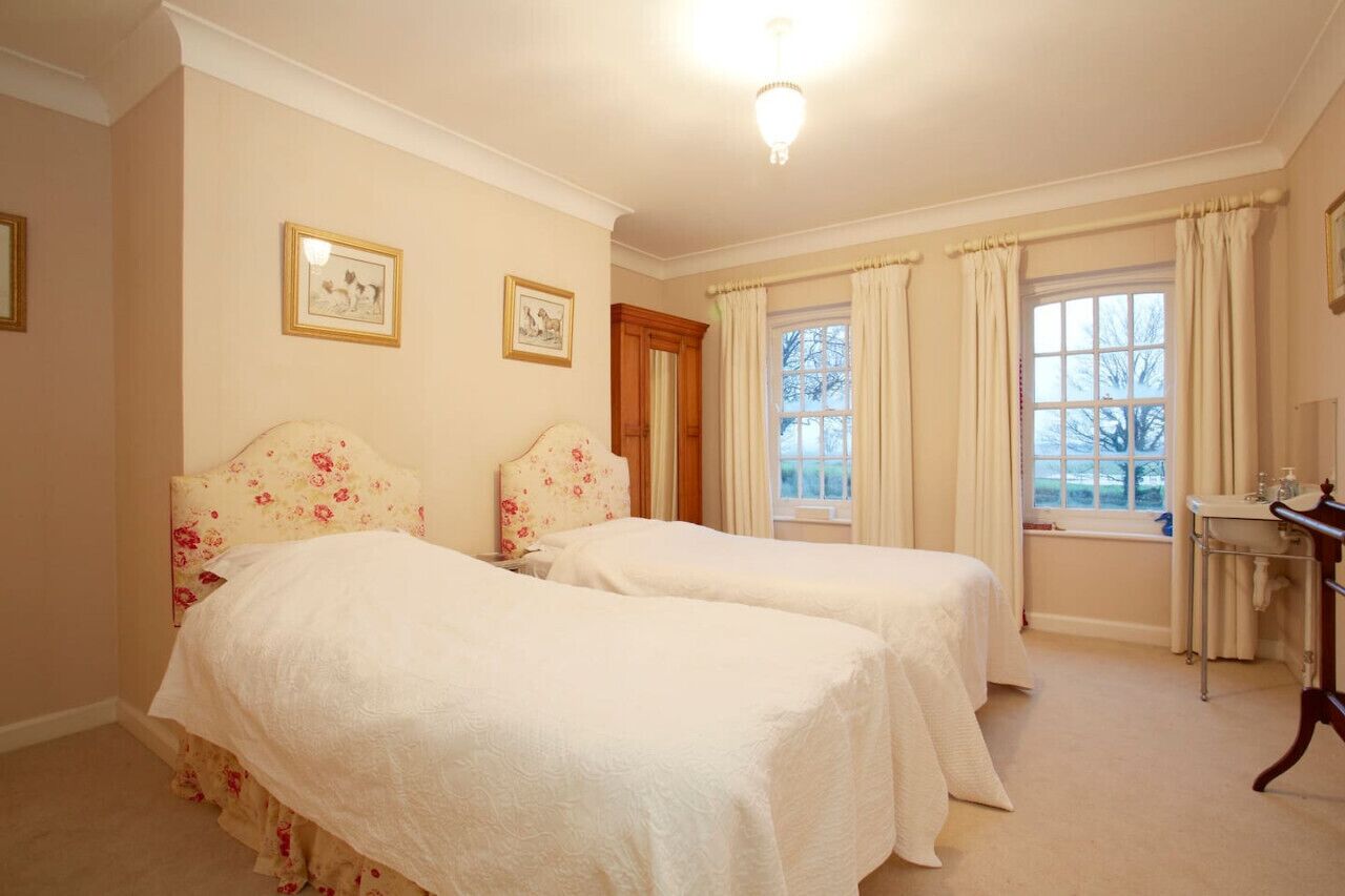 Una habitación con dos camas, una ventana con cortinas, una mesita de noche y cuadros enmarcados en la pared.