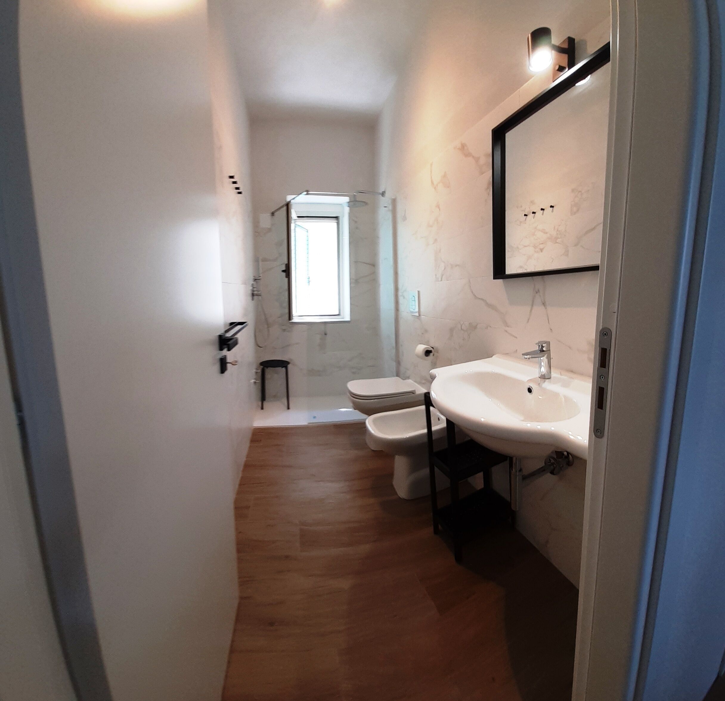 Et aflangt badeværelse med trægulv, toilet, bidet og vask med spejl over.