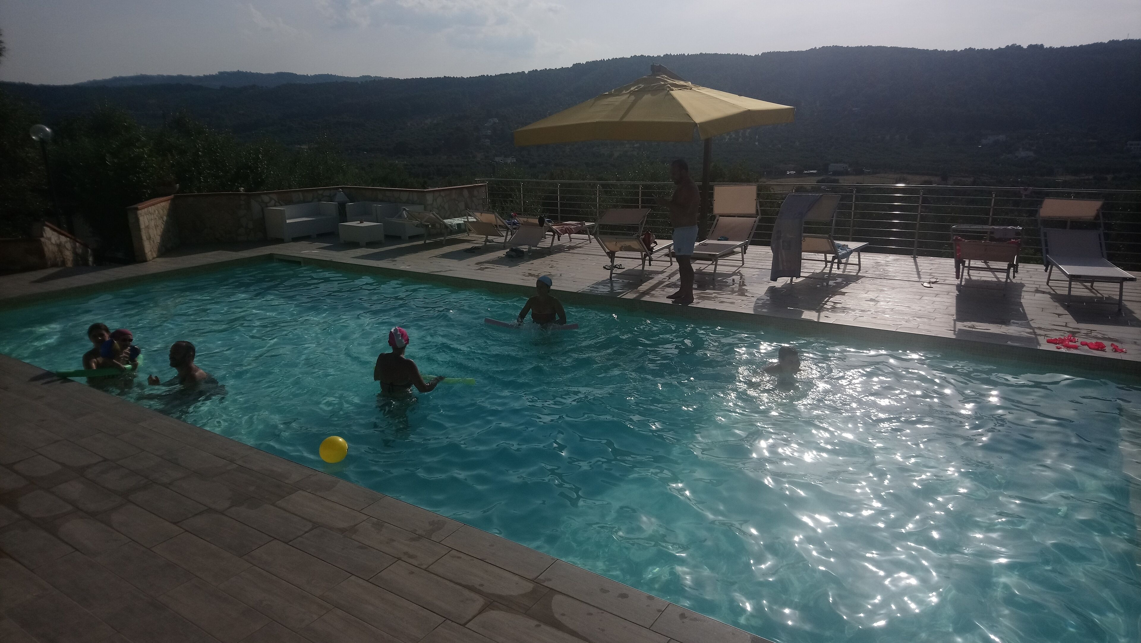 Ein Schwimmbecken mit darin badenden Personen, umgeben von Liegestühlen und einem Sonnenschirm.