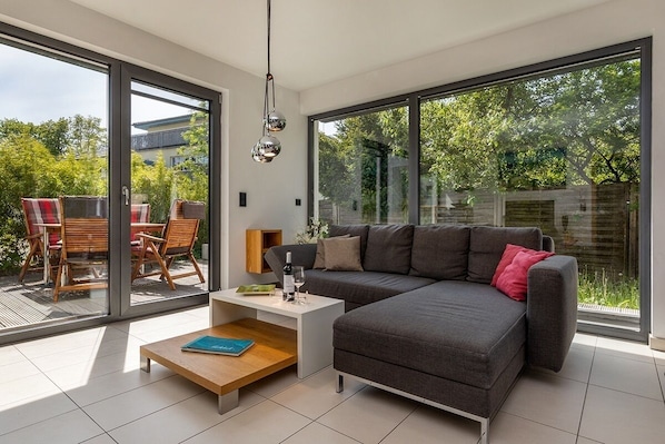 A modern living room with a grey sofa, a wooden coffee table, and a glass door leading to an outdoor patio with wooden furniture.