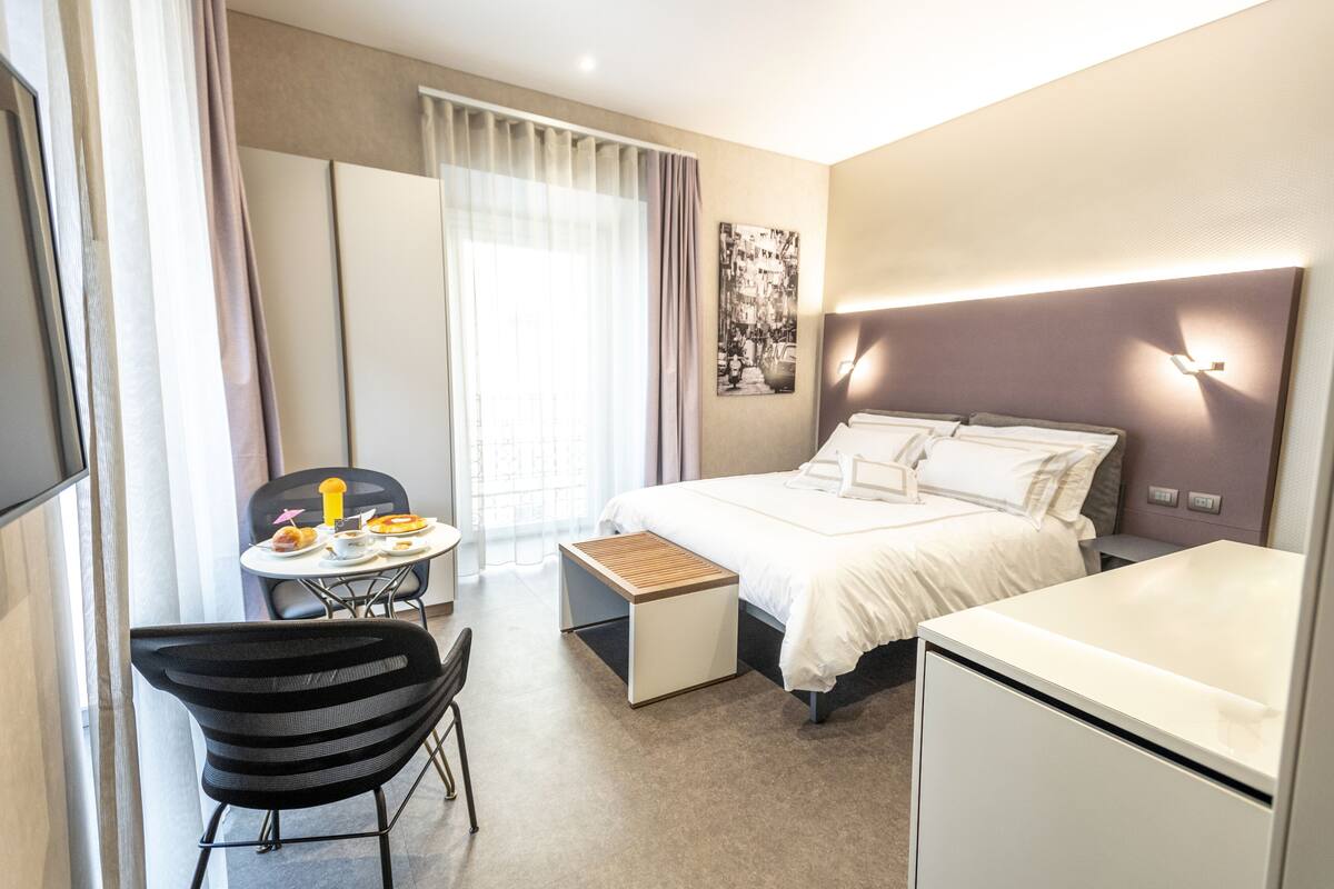 Una habitación de hotel moderna con cama, mesita de noche, silla y una pequeña zona de escritorio.