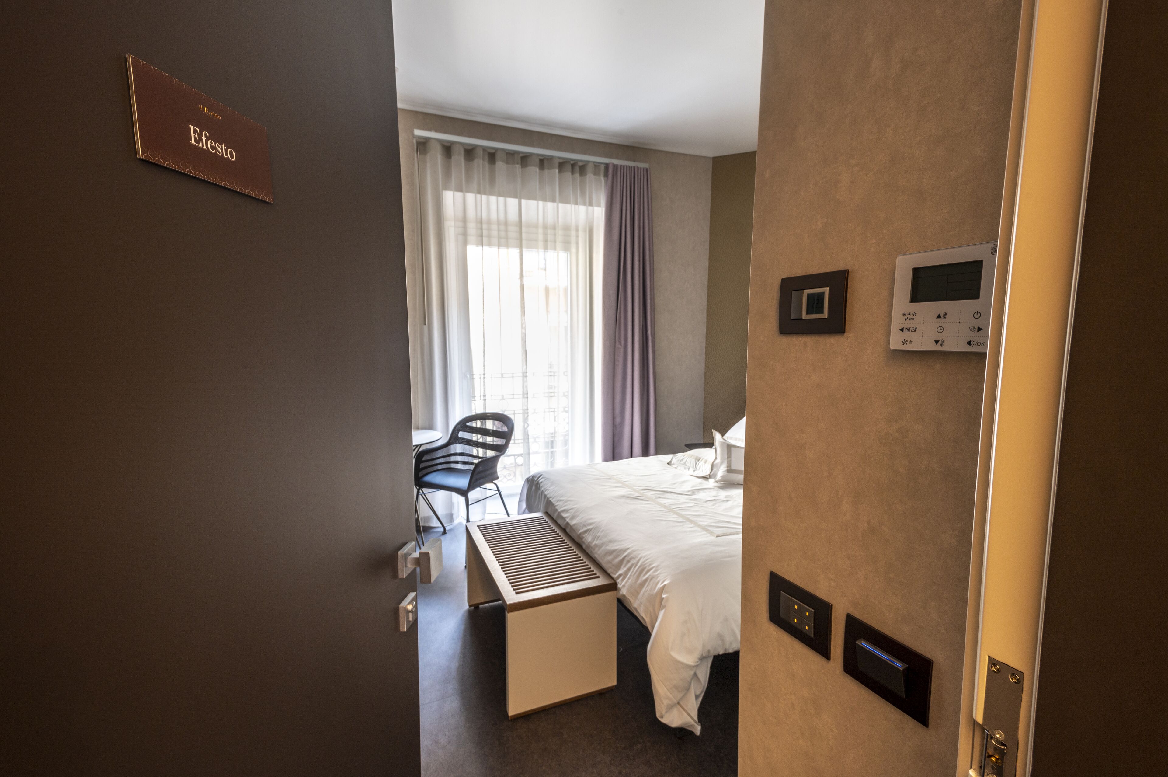 A hotel room with a bed, a desk, a chair, a window with curtains, and a thermostat on the wall.