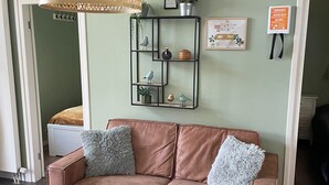 Una sala de estar con un sofá marrón, una mesa de centro redonda de madera, un candelabro colgante de ratán, un estante de metal negro con objetos decorativos, y una pared verde con un cuadro enmarcado.