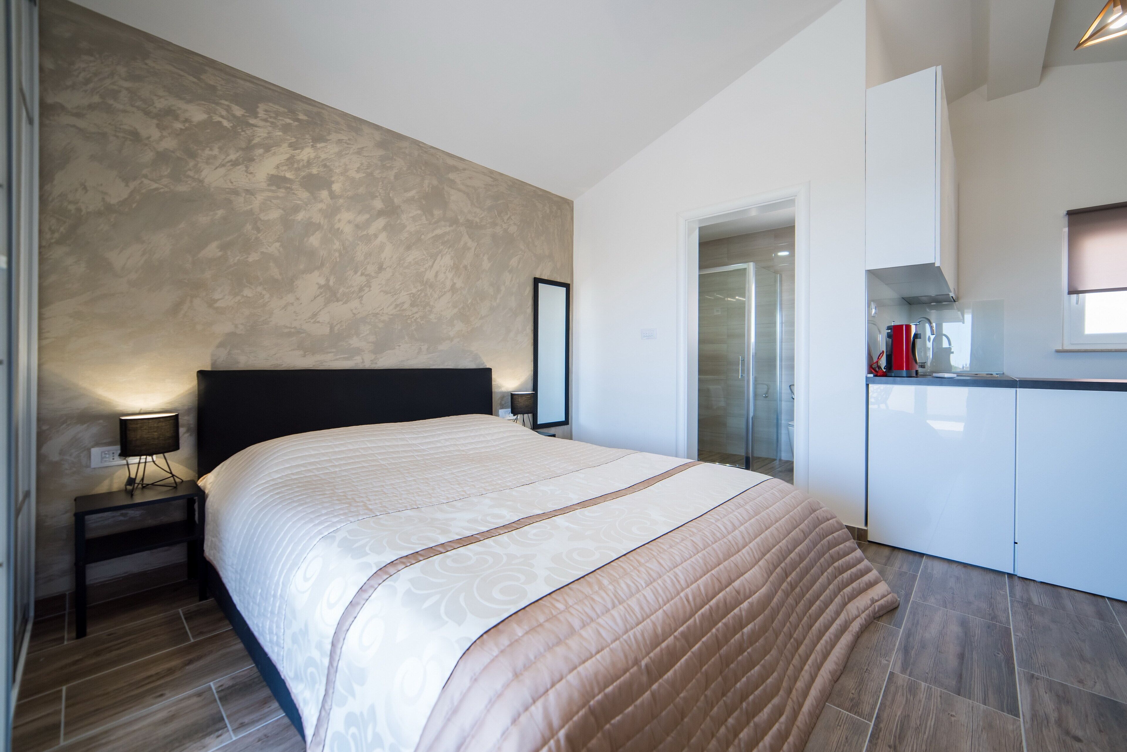 A modern hotel room with a large bed, a bedside table with a lamp, a kitchenette, and a bathroom visible through a glass door.