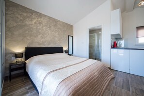 A modern hotel room with a large bed, a bedside table with a lamp, a kitchenette, and a bathroom visible through a glass door.