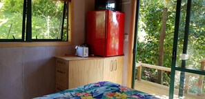 Un dormitorio con cama, un armario de madera, un refrigerador rojo y una ventana con vista a la vegetación.