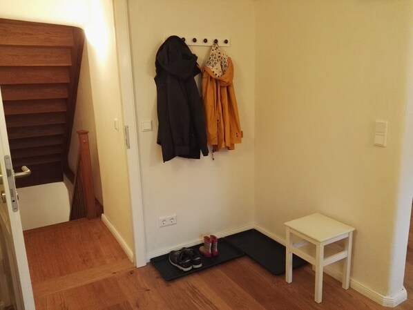 Ein Flur mit einem Kleiderständer, an dem zwei Jacken hängen, einem kleinen weißen Tisch und einer schwarzen Matte auf dem Boden.