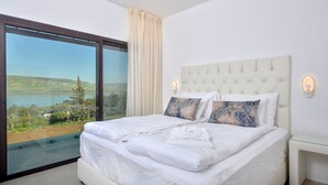 Een slaapkamer met een groot bed, twee kussens, een nachtkastje en uitzicht op een meer en heuvels.