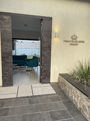 Ein moderner Hotel Eingang mit Blick auf einen See, eine Sitzecke mit türkisfarbenen Stühlen und ein Schild mit der Aufschrift 'PRIMOR DELUXE SUITES KINNERET'.