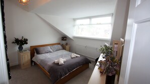 A bedroom with a sloped ceiling, a bed with a patterned bedspread, a bedside table with a lamp, a window with blinds, and a potted plant. - Lymm village apartment (Lymm)